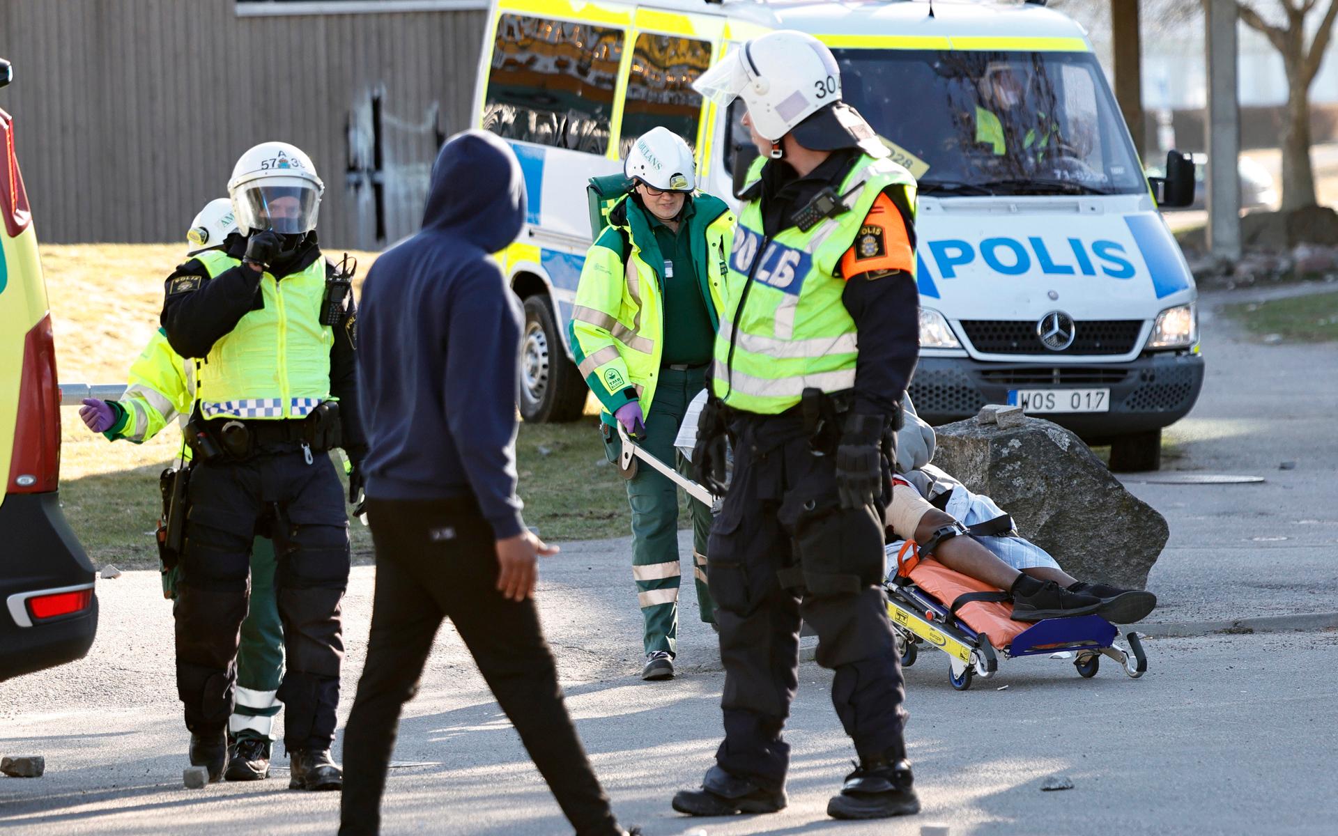 Polis och ambulanspersonal lägger en skadad man som skjutits i benet på en bår under upploppen i Navestad i Norrköping på påskdagen.Oroligheterna utlöstes av att den högerextrema provokatören Rasmus Paludan har sagt att han ska återvända till Östergötland för nya demonstrationer under dagen.
